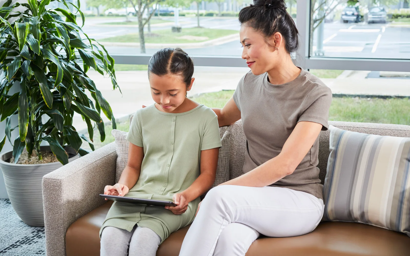 Adult and child seated on a striped upholstered sofa while looking at a tablet beside a window