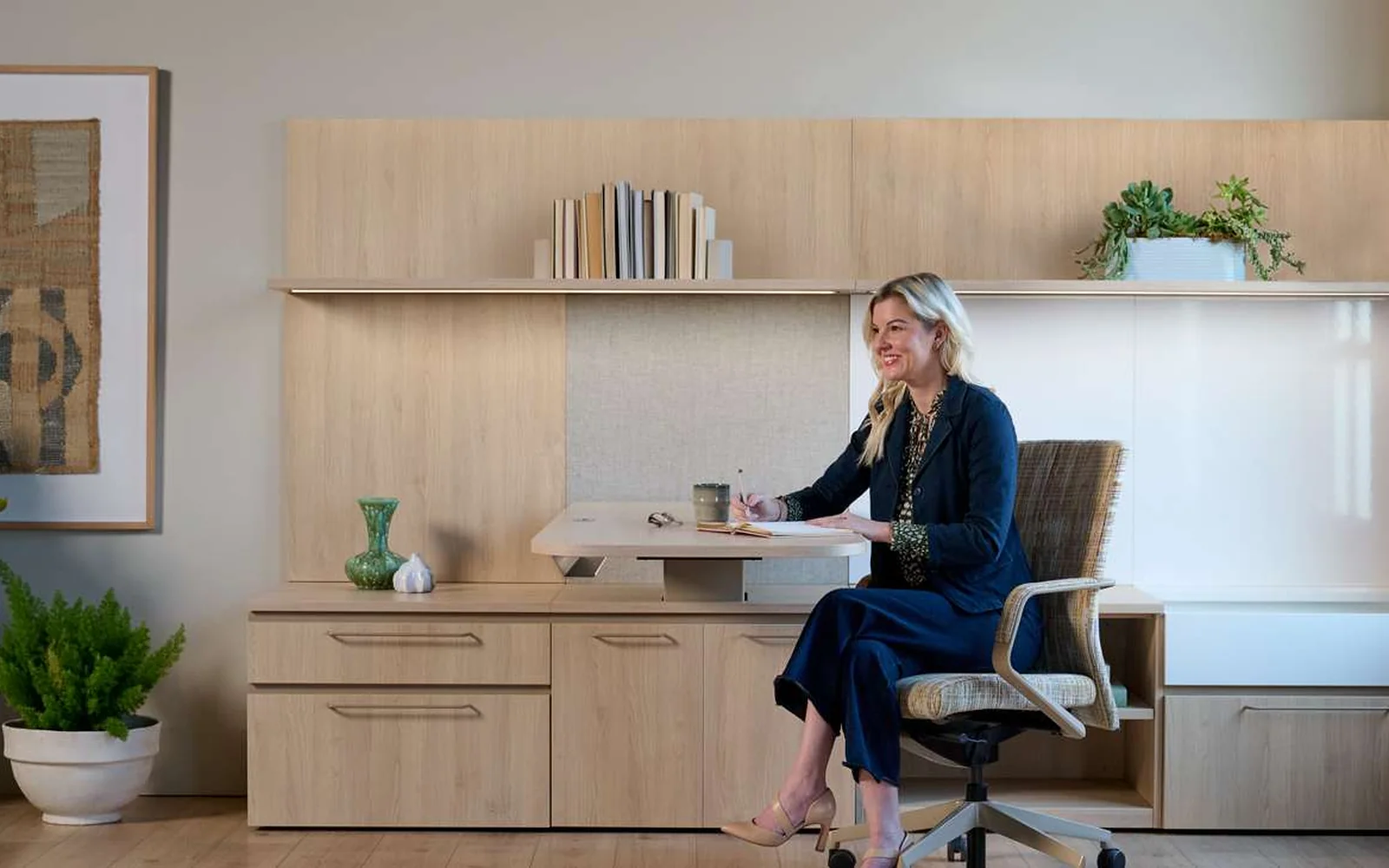 OFS Adler private office with a woman at a light-wood desk, integrated wall storage, shelving, plants, and an exposed brick wall
