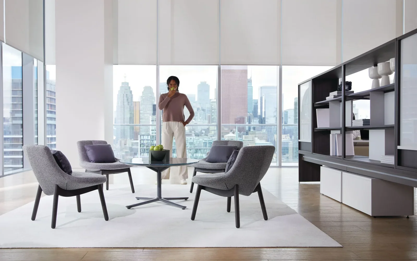 Teknion private-office setting with four gray lounge chairs, a small table, wall storage, and a woman beside skyline windows